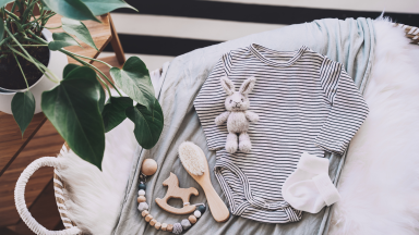 Still life background of cute baby products - changing basket with baby bodysuit, newborn clothes, knitted rabbit and wooden toy