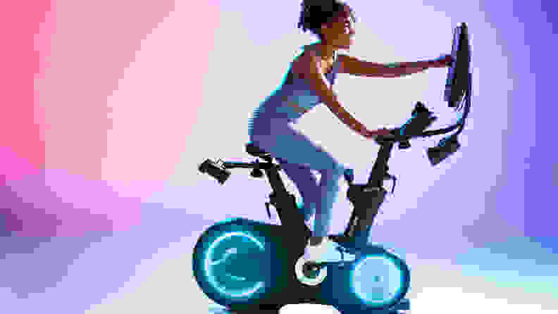 Woman riding the Echelon EX-8s Exercise Bike against a pink and purple background.