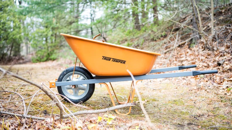 An orange True Temper wheelbarrow sits in a wooded area.