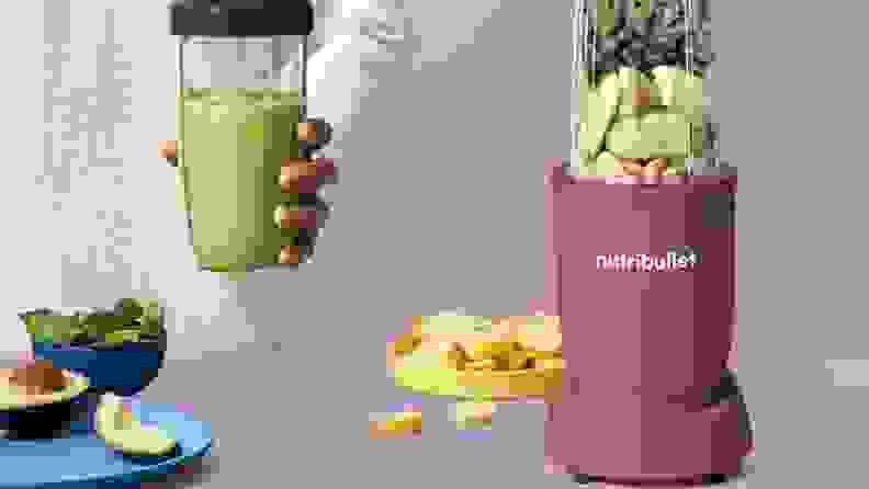 A pastel purple personal blender filled with fruit next to a hand holding a blended green smoothie.