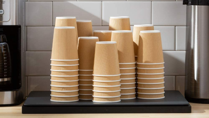 There are many paper coffee cups stacked on top of an Amazon Dash Smart Shelf. To the left of the shelf is a coffee maker and to the right appears to be a hot water dispenser.