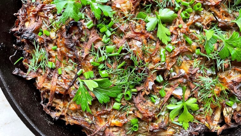 A closeup photo of crispy potato kugel in a cast-iron skillet topped with dill, parsley, and chives.