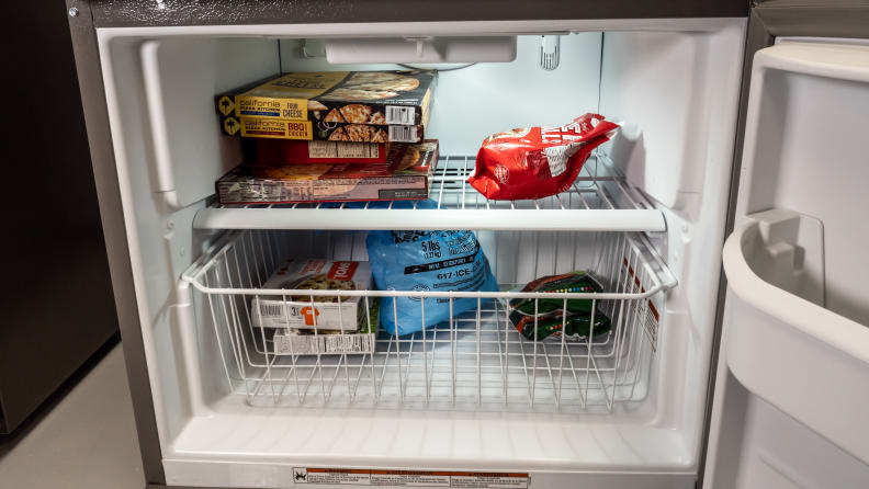 A close-up of the freezer. Inside is a single wire latticework shelf, above a basket  made from the same kind of wire. The drawer and shelf are stocked with frozen foods.