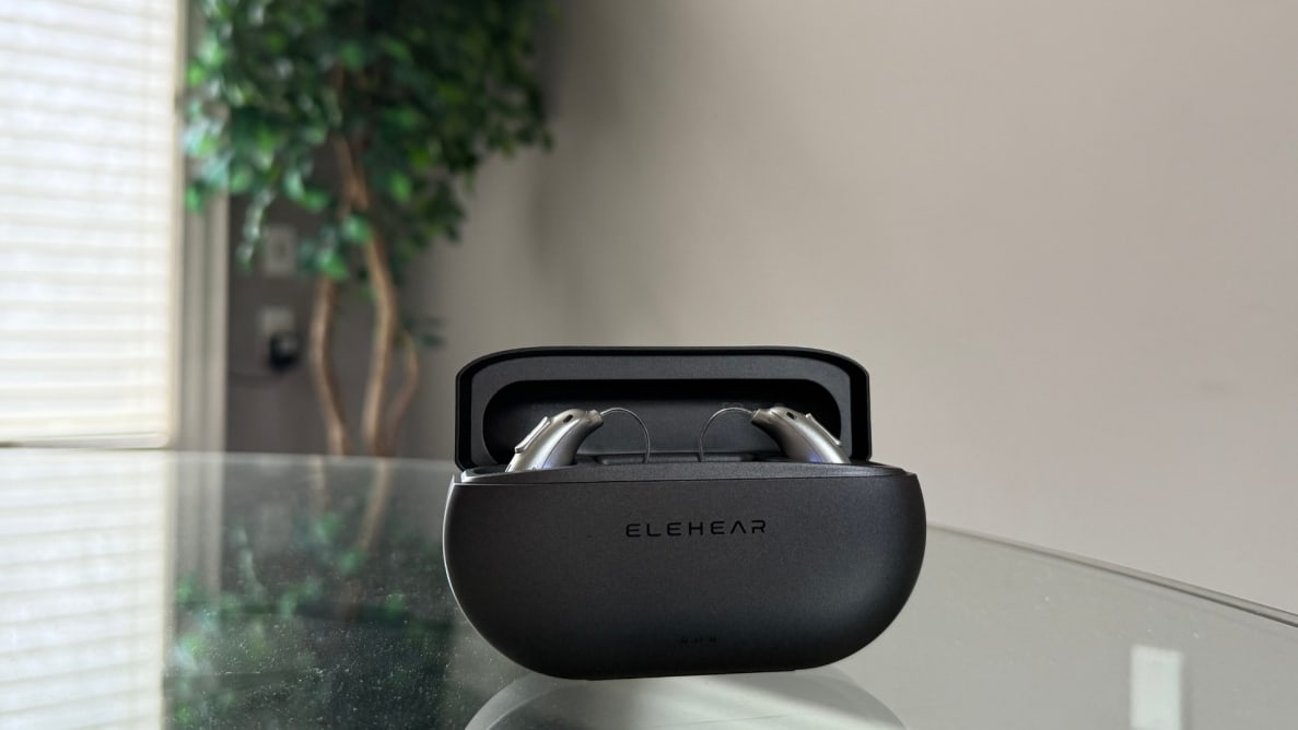 An open black charging case with hearing aids inside sits on top of a glass table with a tree in the background