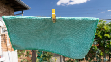 Teal microfiber cloth being hung on clothesline by one one yellow clip outdoors on a sunny day.