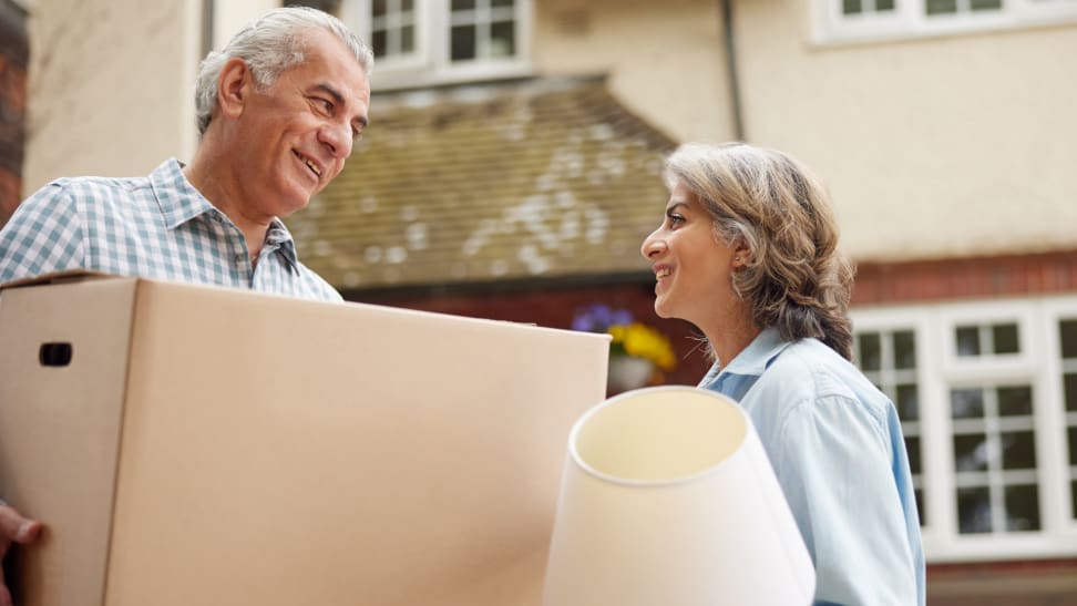 Two people moving into a house stand outside carrying a box and a lamp.