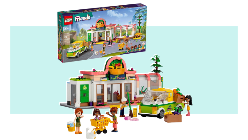 The pieces of the Fresh Market organic grocery store Lego Friends set, including the store, shoppers, a worker sweeping, and a worker loading a car, stand in front of the toy package.