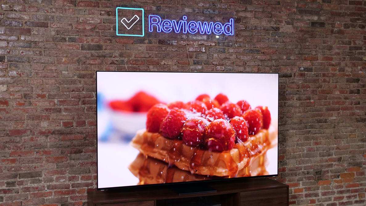 The Samsung S95C QD-OLED TV on a entertainment center and Reviewed sign about the TV, with a brick wall in the background.