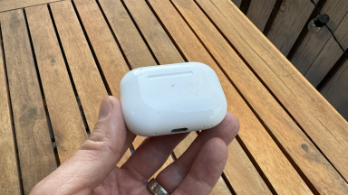 A hand holding the AirPods Pro 2 charging case with USB-C above a wooden table outside.