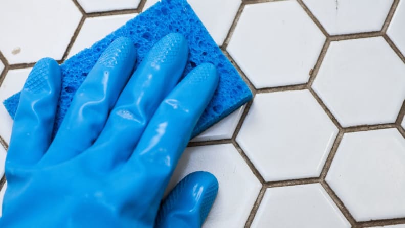 Hand wearing blue rubber gloves and blue sponge to wipe tile surface.