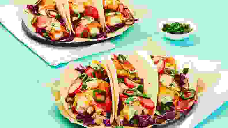 Three fish tacos arranged on a square white plate, accompanied by a second plate of tacos in the background.