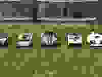 Five robot lawn mowers lined up together in the green grass with a fence behind