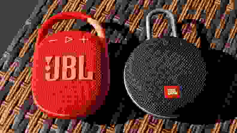 The oval shaped, brilliantly red JBL Clip 4 sits next to circular, black Clip 3 on a bar made of criss-crossed brown and black vinyl.