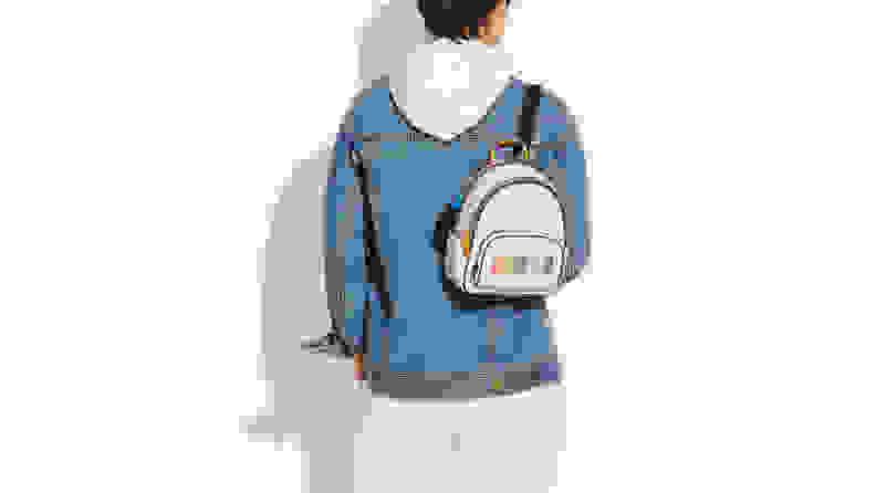 Person facing away wearing denim jacket, white pants, and white hoodie under jacket, and a white and black mini backpack that has "COACH" written across it in rainbow colors