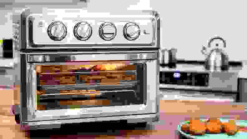 An image of a Cuisinart Air Fryer and Toaster oven seated on a tabletop. The viewer can see food within the air fryer on the metal tray inside.