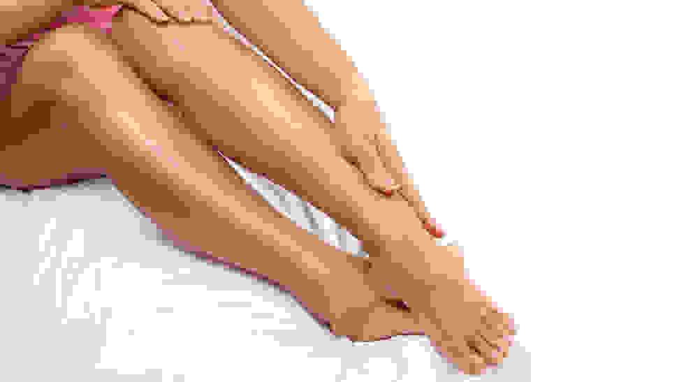 A person sits on a bed with their legs stretched out and touches their ankle with their hand.