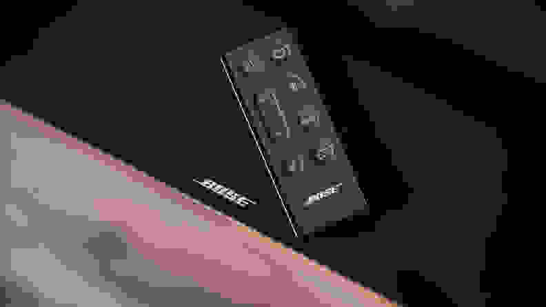 The Bose TV Speaker remote resting on top of the all-black soundbar, the white Bose logo peaking out beneath the slim rectangular remote with bubbled buttons on top.