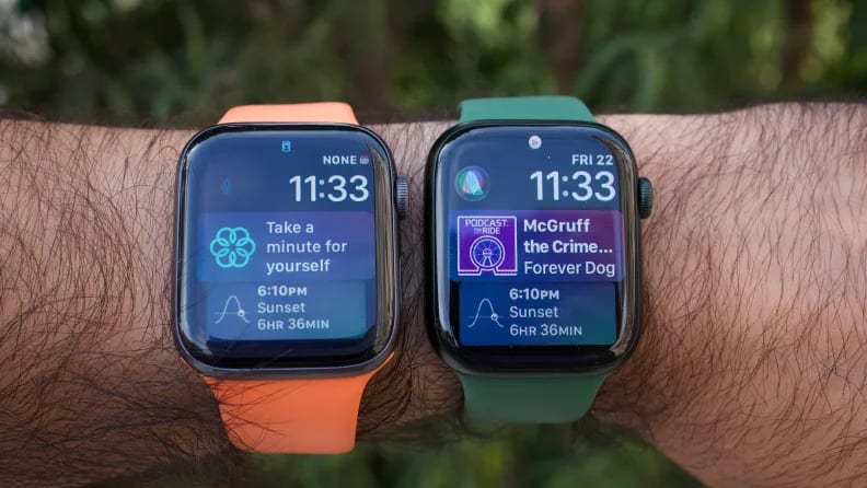The Apple Watch Series 5 (which shares the same size display as the SE) next to the Apple Watch Series 7, both being worn on a wrist, showing Apple's Siri watchface.