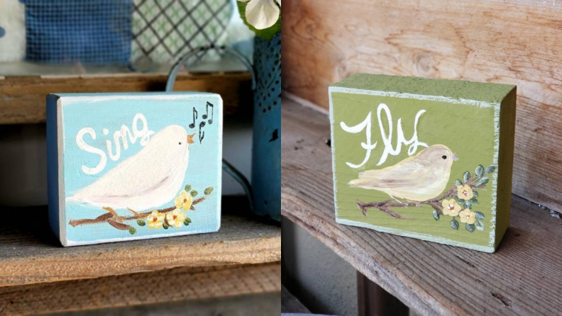 On left, blue wooden garden sign with hand painted white bird on front that reads "Sing."  On right, wooden garden sign with hand painted white bird on front that reads "Fly."