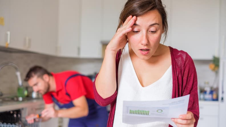 Woman looking at repair bill while repairman works on dishwasher in background