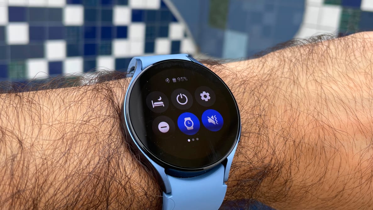 The Galaxy Watch 5 sits on a wrist with a blue band in front of a tiled background.