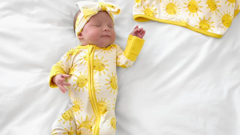 A sleeping baby wearing a yellow sun-print onesie and matching headband lies on a white bed.