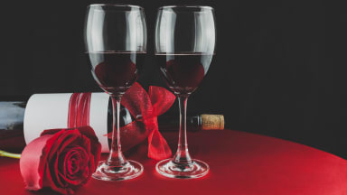 A romantic setting featuring two glasses of red wine on a red tablecloth, accompanied by a bottle of wine wrapped in a glittery red bow and a single red rose, with an "Editors' Choice Reviewed" badge in the top right corner.