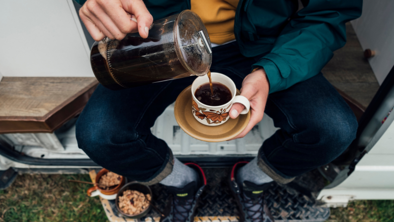 A person is pouring coffee from a French press into. a mug while sitting in a camper van.