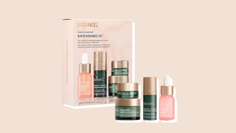 A collection of four Biossance Rapid Radiance skincare products sit stacked in front of their box. They're in green, gold, and pink-colored containers.