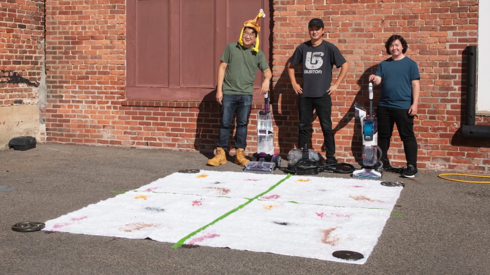 Three people stand in front of a brick wall and next to a white rug with stains on it.