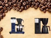 Two coffee pots sit on a beige background surrounded by coffee beans