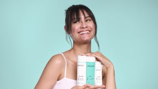 A smiling young woman with acne holds up three proactiv acne treatment products against a teal background.
