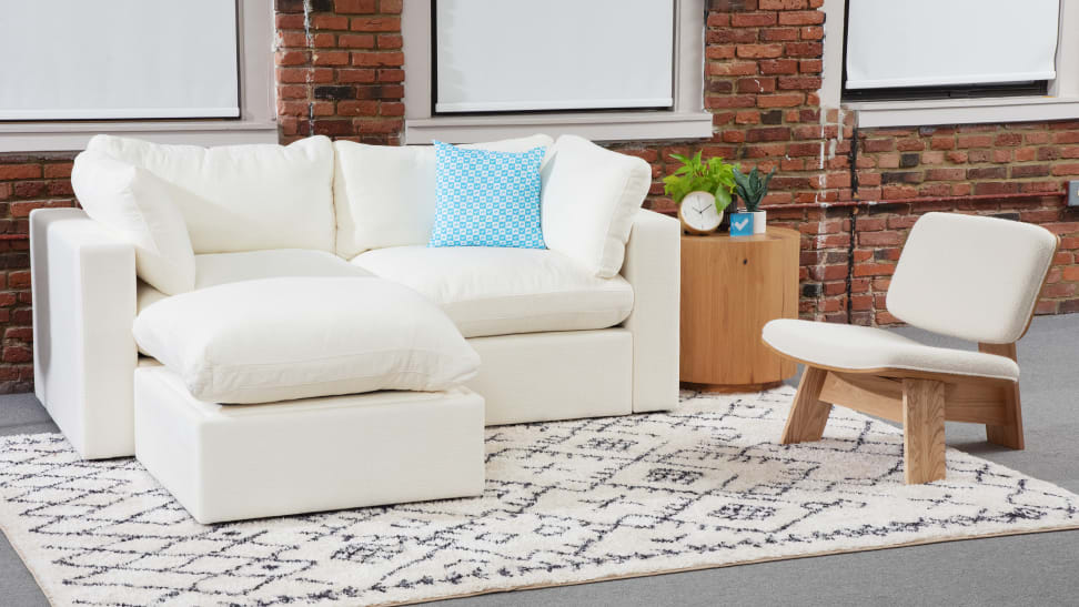 The Cozey couch show in white with a white and wood accent chair next to it, staged against a brick wall.