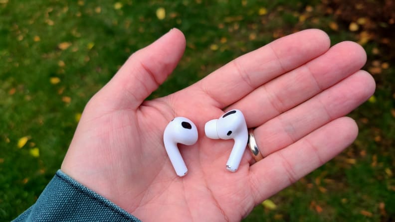 The all-white AirPods (left) next to the all-white AirPods Pro (right) shows similar sizing in the reviewer's hand, but the pro shows a silicone eartip.