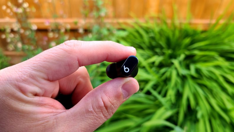 The black Beats Studio Bud is held with its dial knob facing forward in front of a leafy garden.