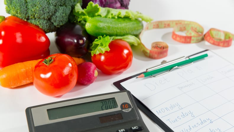 A calculator sitting next to a clipboard with diet tracking weekly schedule on paper in front of an array of colorful vegetables.