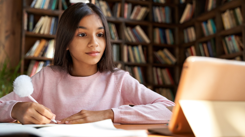 A young child writes at a desk while watching a tutorial.