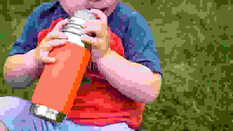 A child sitting on the grass holds a stainless steel Pura Kiki straw cup with an orange sleeve.