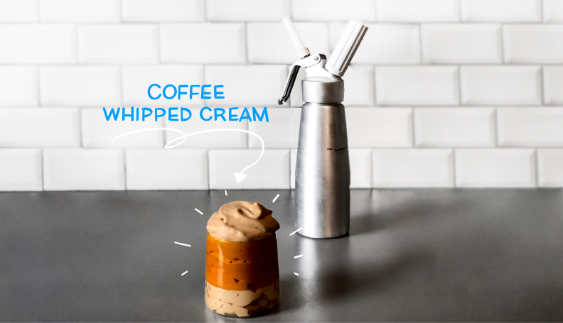 An iced coffee beverage topped with coffee whipped cream sits on a kitchen counter with a whipped cream canister in the background.