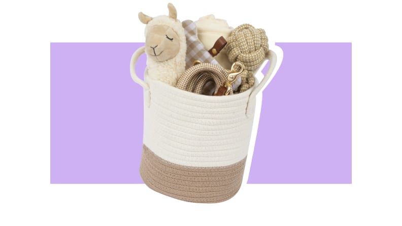 A knit white and basket with a brown bottom rim containing a llama toy, treats, and other products against a purple background.