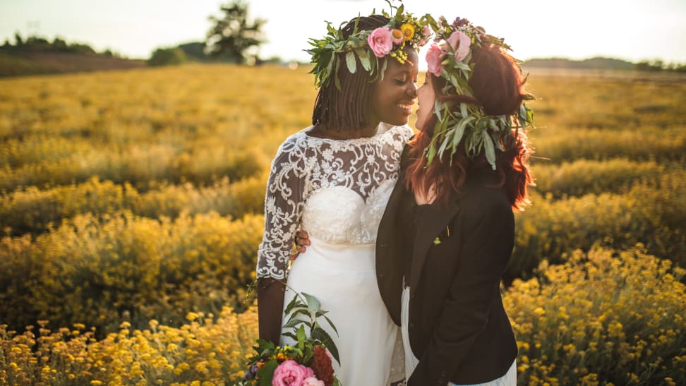 A Black human in a wedding dress and white human in a suit celebrate their wedding day, leaning in for a kiss.