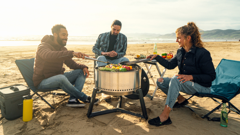 Three people grill vegetables on a Solo Stove while camping on a beach.