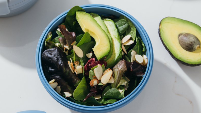 In the center of the image, there's a Stojo bowl filled with salad with sliced avocado and almonds on top. A half avocado is next to it.