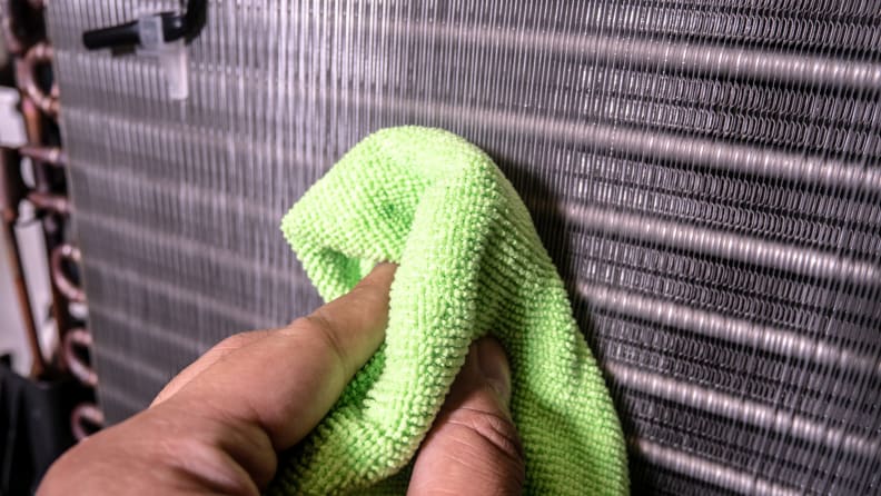 A hand reaches into frame to clean the grill on the back of the A/C with a cloth.