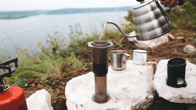 A person is pouring water over an Aeropress coffee maker at a riverbank.