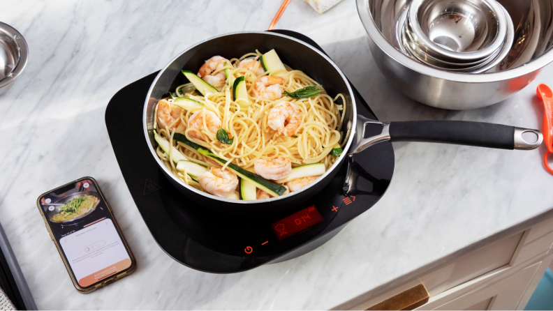 A bowl of shrimp noodles with a smartphone displaying the Tramontina app next to it.