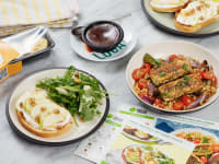 Four plates of HelloFresh meals, a package of egg bites, and recipe cards spread out on a counter.