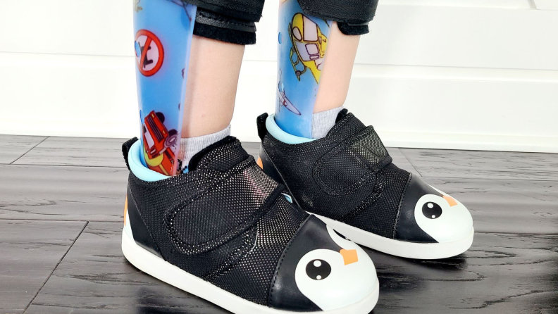 Small child's legs and feet displaying colorful leg braces and the Ikiki Squeaky penguin shoe.