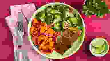 Plate filled with carrots, broccoli and salmon next to small plant, cutlery and limes on top of table.