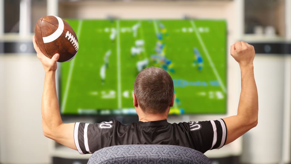 The back of a person's head as they celebrate with a football in front of a very large TV displaying an American football game